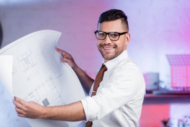 Gözlüklü mutlu mimar elinde planla kameraya gülümsüyor.