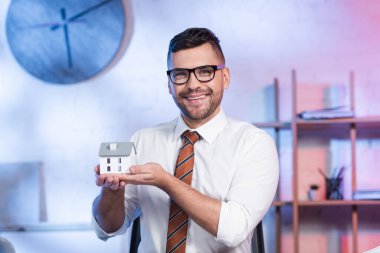 Gözlüklü neşeli mimar ev modelini tutuyor ve kameraya gülümsüyor.