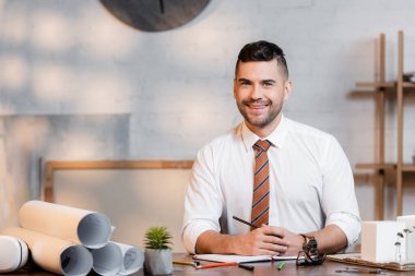 smiling architect looking at camera while sitting at workplace near rolled blueprints clipart