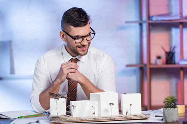 улыбающийся архитектор сидит на рабочем месте рядом с моделями домов