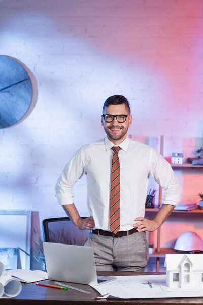 happy architect standing with hands on hips near laptop and house model on desk