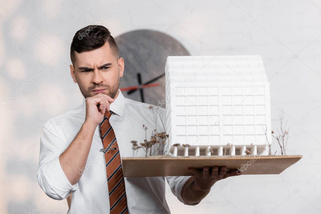 thoughtful architect with house maquette holding hand near chin in office