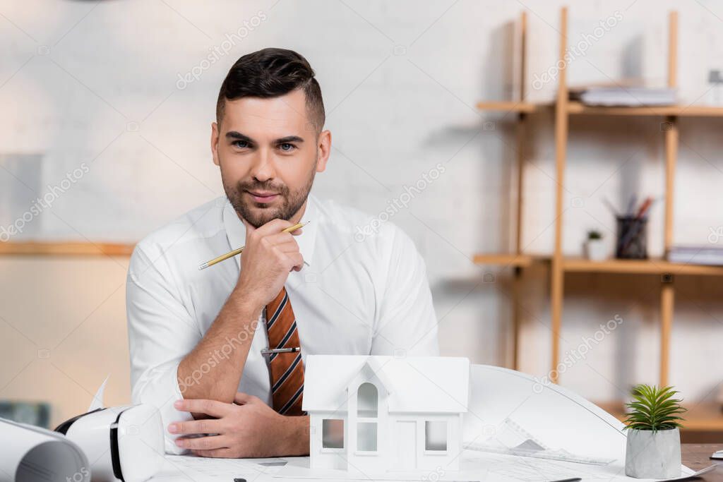 smiling architect looking at camera near house model and blueprints on desk