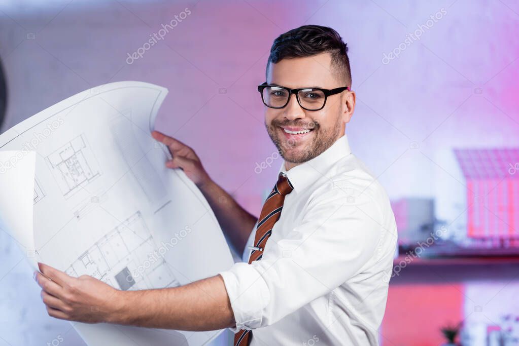 happy architect in eyeglasses smiling at camera while holding blueprint