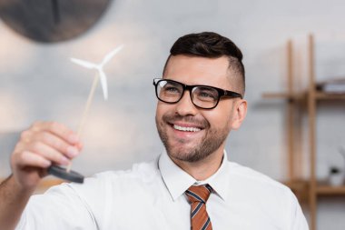 cheerful architect in eyeglasses smiling while holding model of wind turbine clipart