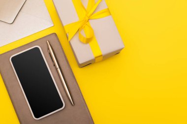 Zarfın yanında kalem ve sarı üzerine izole edilmiş hediye kutusu olan boş ekranlı akıllı telefonun üst görünümü