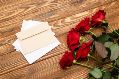 Zarfların yanında kırmızı güllerin yüksek açılı görüntüsü ve ahşap yüzeyde kağıtlar 