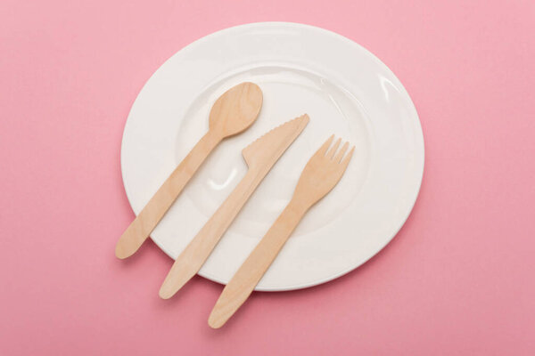 top view of disposable wooden cutlery on white plate isolated on pink 