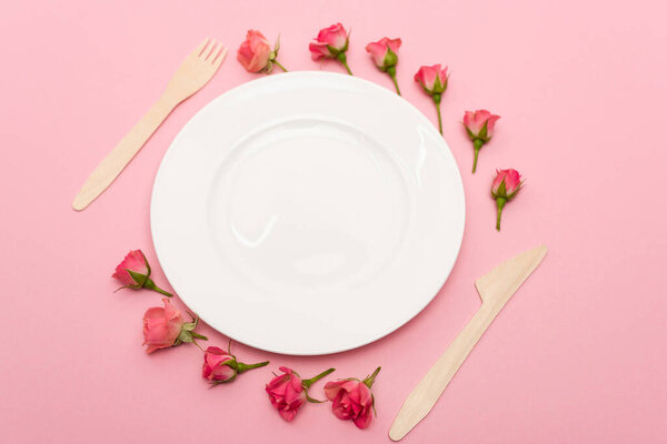 high angle view of disposable wooden cutlery near white plate and flowers isolated on pink 