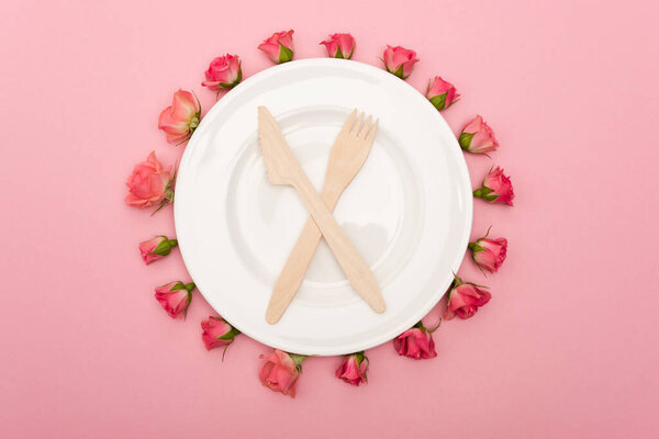 top view of disposable wooden cutlery on white plate and tea roses isolated on pink 