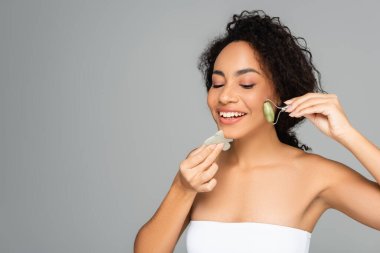 Young african american woman smiling while using jade roller and gua sha isolated on grey  clipart