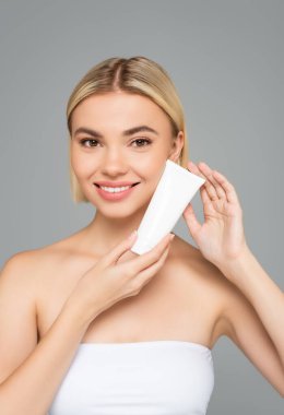 Neşeli kadın kameraya bakıyor ve elinde kozmetik kremiyle gri renkli bir tüp tutuyor. 