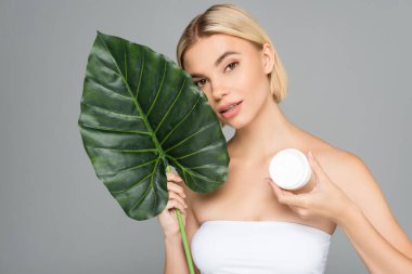 Kozmetik kremi ve tropikal yaprağı olan bir kadın gri renkli kameraya bakıyor. 