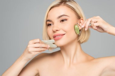 Blonde woman with clean skin massaging face with gua sha and jade roller isolated on grey  clipart