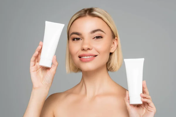 Smiling blonde woman holding tubes with cosmetic cream isolated on grey