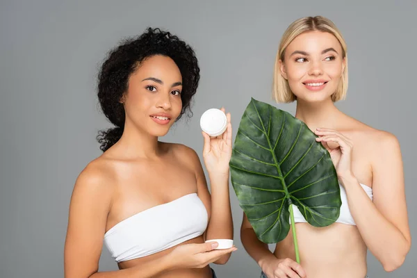 Smiling multiethnic women holding cosmetic cream and topical leaf isolated on grey