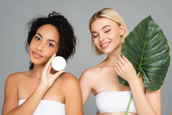 Sorridente Donne Interrazziale Posa Con Crema Cosmetica Foglia Tropicale Isolato — Foto Stock