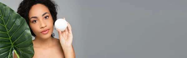 African american woman holding cosmetic cream and green leaf isolated on grey, banner