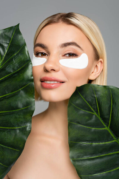 Молодая женщина с идеальной кожей и повязками на глазу позирует рядом с тропическими листьями, изолированными на сером 