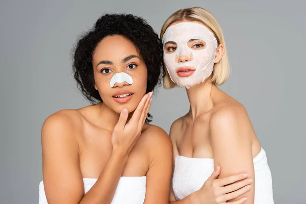 Multiethnic women with cleansing stripe and sheet mask looking at camera isolated on grey