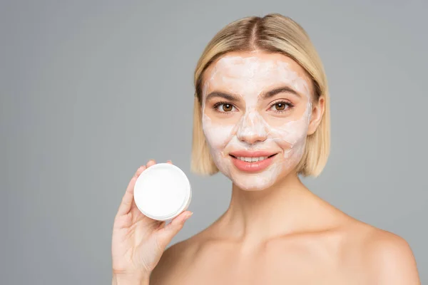 Young woman with naked shoulders holding cosmetic cream isolated on grey