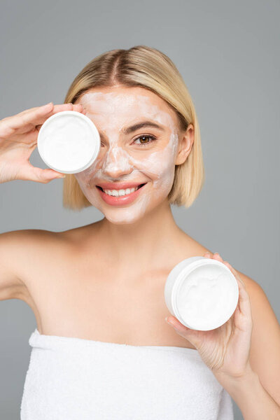 Веселая женщина в полотенце и маске для лица с косметическим кремом, изолированным на сером 