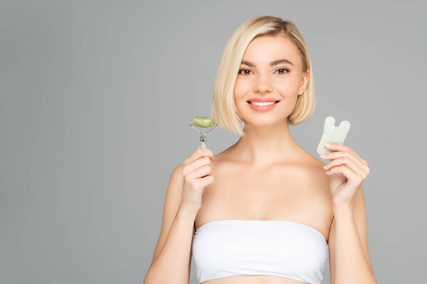 Smiling blonde woman holding gua sha and jade roller isolated on grey 