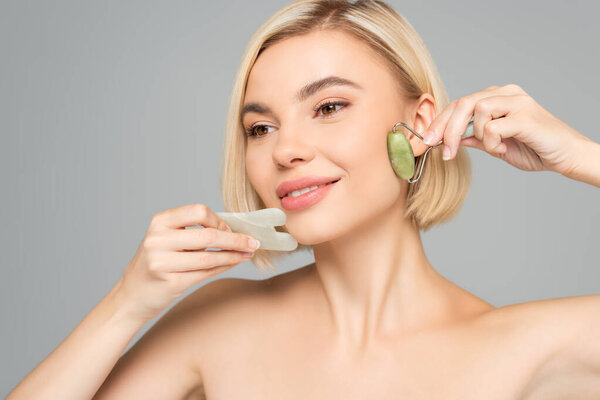 Blonde woman with clean skin massaging face with gua sha and jade roller isolated on grey 