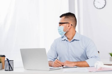 Medikal maskeli ve gözlüklü işadamı elinde dizüstü bilgisayarla kalem tutarken gözünü kaçırıyor.