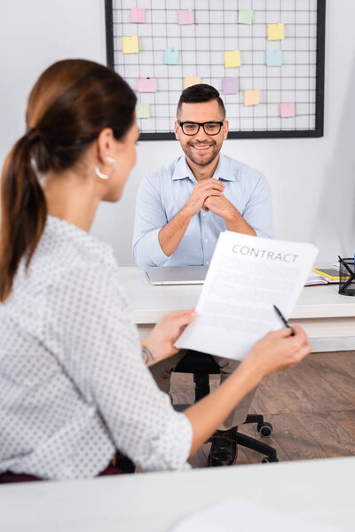 счастливый бизнесмен в очках глядя на деловую женщину, заключившую контракт на размытом переднем плане 