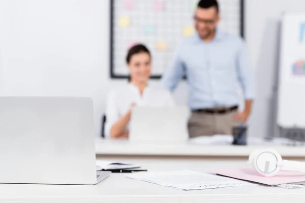 laptop near papers and headphones on desk near business people on blurred background