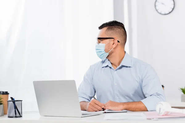 Üzletember Orvosi Maszkban Szemüvegben Félrenéz Miközben Tollat Tart Notebook Laptop — Stock Fotó