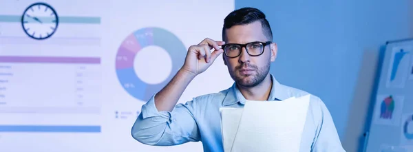 Hombre Negocios Ajustando Gafas Sosteniendo Papeles Cerca Gráficos Gráficos Pared —  Fotos de Stock