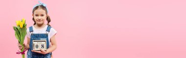 Elinde lale ve tahta küplerle mutlu bir çocuk. Pembe pankartta 8 Mart yazıları var.