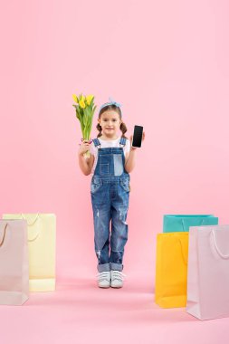 Elinde boş ekranlı akıllı telefon ve pembe çantaların yanında laleler tutan bir çocuk.