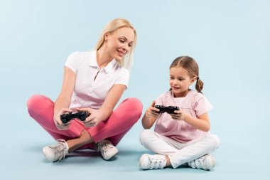 KYIV, UKRAINE - 25 Kasım 2020: mutlu anne ve kız joystick tutuyor ve bacak bacak bacak üstüne atarken mavi