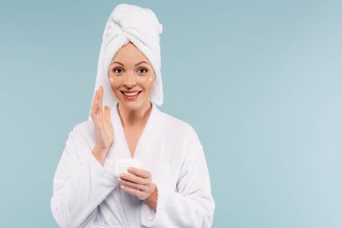 Beyaz bornozlu mutlu kadın elinde yüz kremi kabı ile mavi üzerine izole edilmiş.