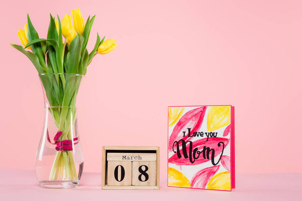wooden calendar with 8 march lettering near greeting card and vase with flowers on pink