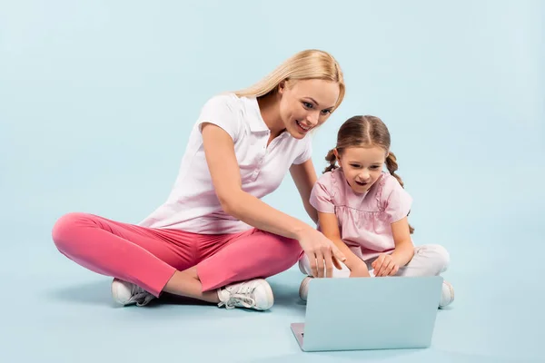 smiling mother and daughter sitting with crossed legs and pointing with finger laptop on blue