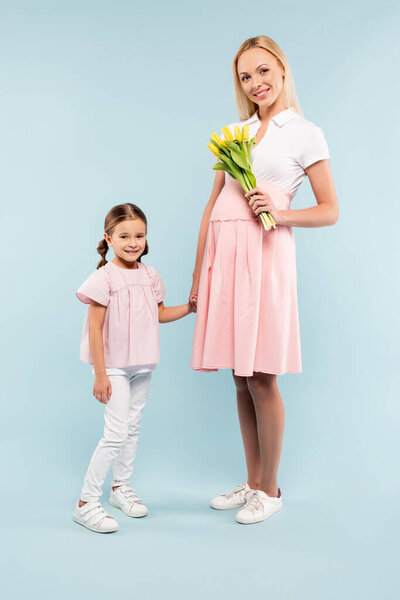 full length of kid holding hands with pregnant mother with tulips on blue