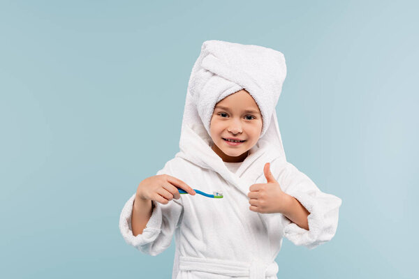 happy kid in bathrobe and towel on head holding toothbrush with toothpaste while showing thumb up isolated on blue