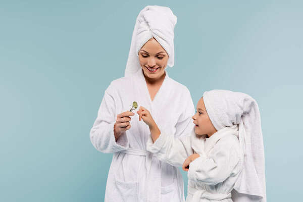 happy kid in bathrobe looking at mother holding jade roller isolated on blue