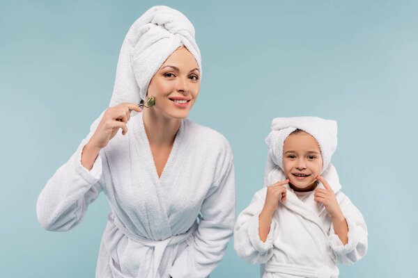 happy kid in bathrobe smiling near mother using jade roller isolated on blue