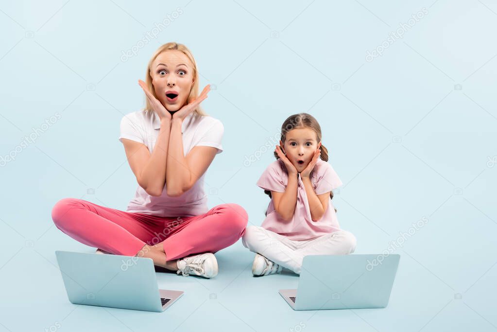 shocked mother and daughter sitting with crossed legs near laptops on blue