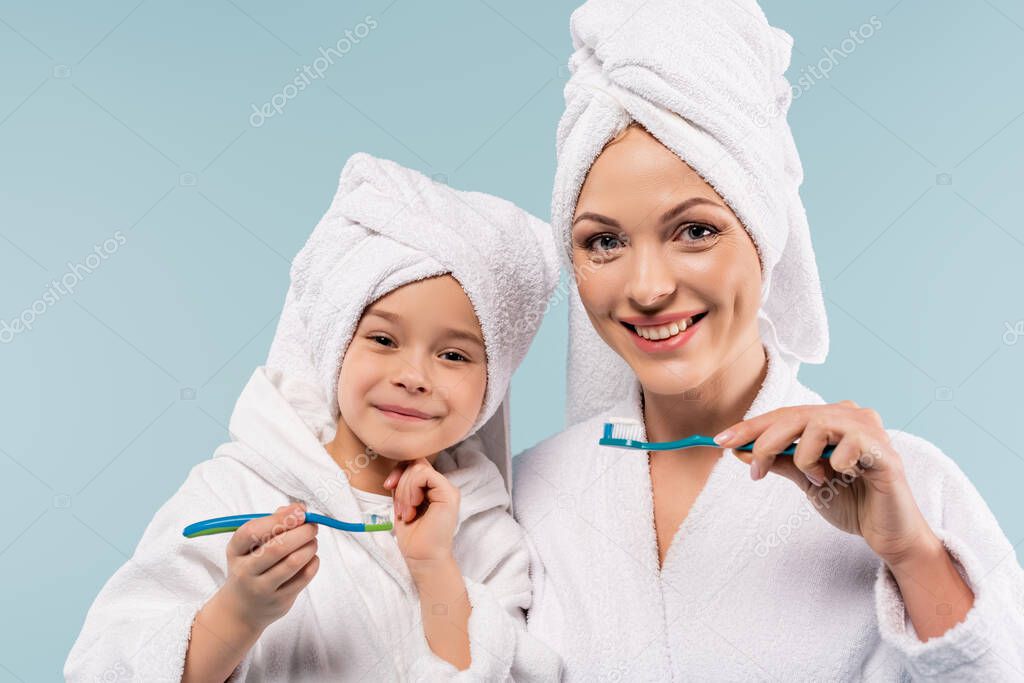 cheerful mother and kid in bathrobes holding toothbrushes with toothpaste isolated on blue
