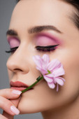 zblízka smyslné ženy s růžovými očními stíny a zavřenýma očima držící květinu izolovanou na šedé