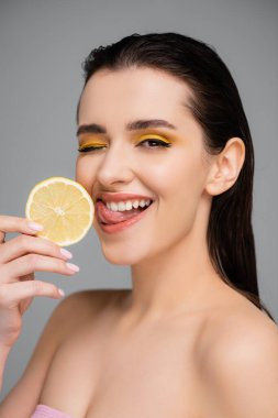 Çıplak omuzlu genç bir kadın dilimlenmiş limon tutuyor ve gri üzerine izole edilmiş bir dil uzatıyor. 
