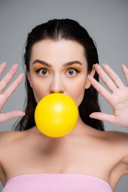 young brunette woman with makeup and bare shoulders blowing yellow bubblegum isolated on grey clipart