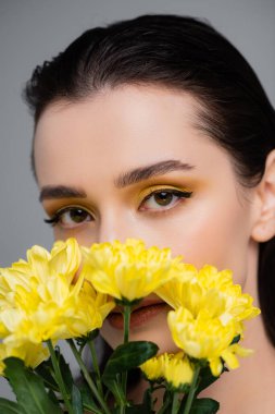 young woman with yellow eyeshadows covering face with blooming flowers isolated on grey clipart