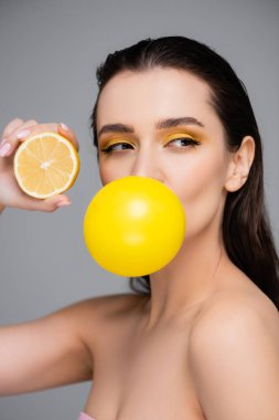 Makyajlı genç bir kadın sarı sakız üflüyor ve limonun yarısını gri renkte tutuyor.